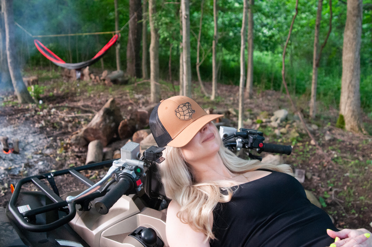 Woman wearing a tan trucker hat with a bear logo patch, sitting on a four-wheeler in a forest camping setting.