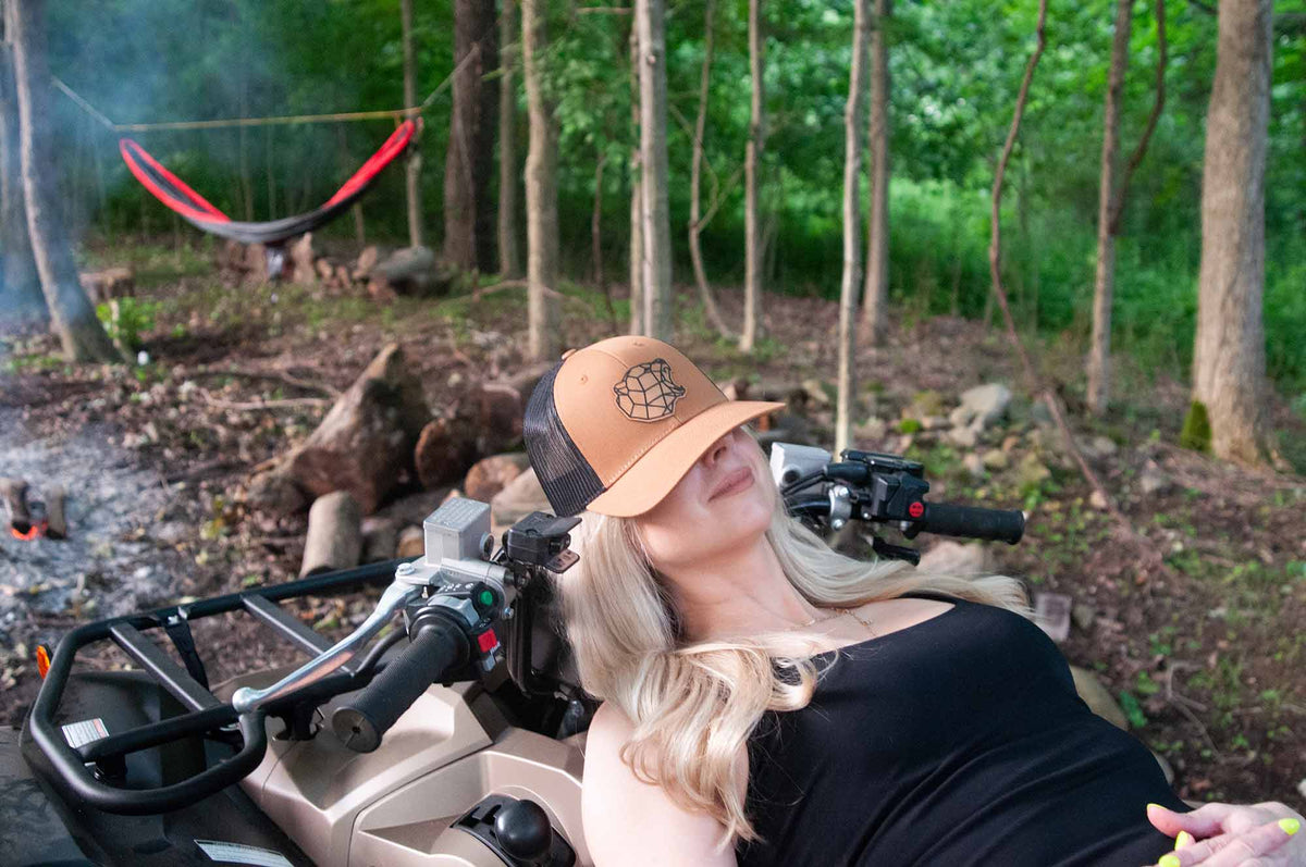 Woman wearing a tan trucker hat with a bear logo patch, sitting on a four-wheeler ATV in a forest camping setting.