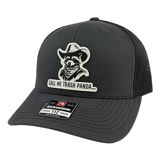 An Artora Design charcoal trucker hat with a black mesh back, featuring a white leatherette laser engraved patch of a western raccoon wearing a cowboy hat and bandana with the text 'CALL ME TRASH PANDA' on the front. A Richardson 112 brand sticker is visible on the brim.